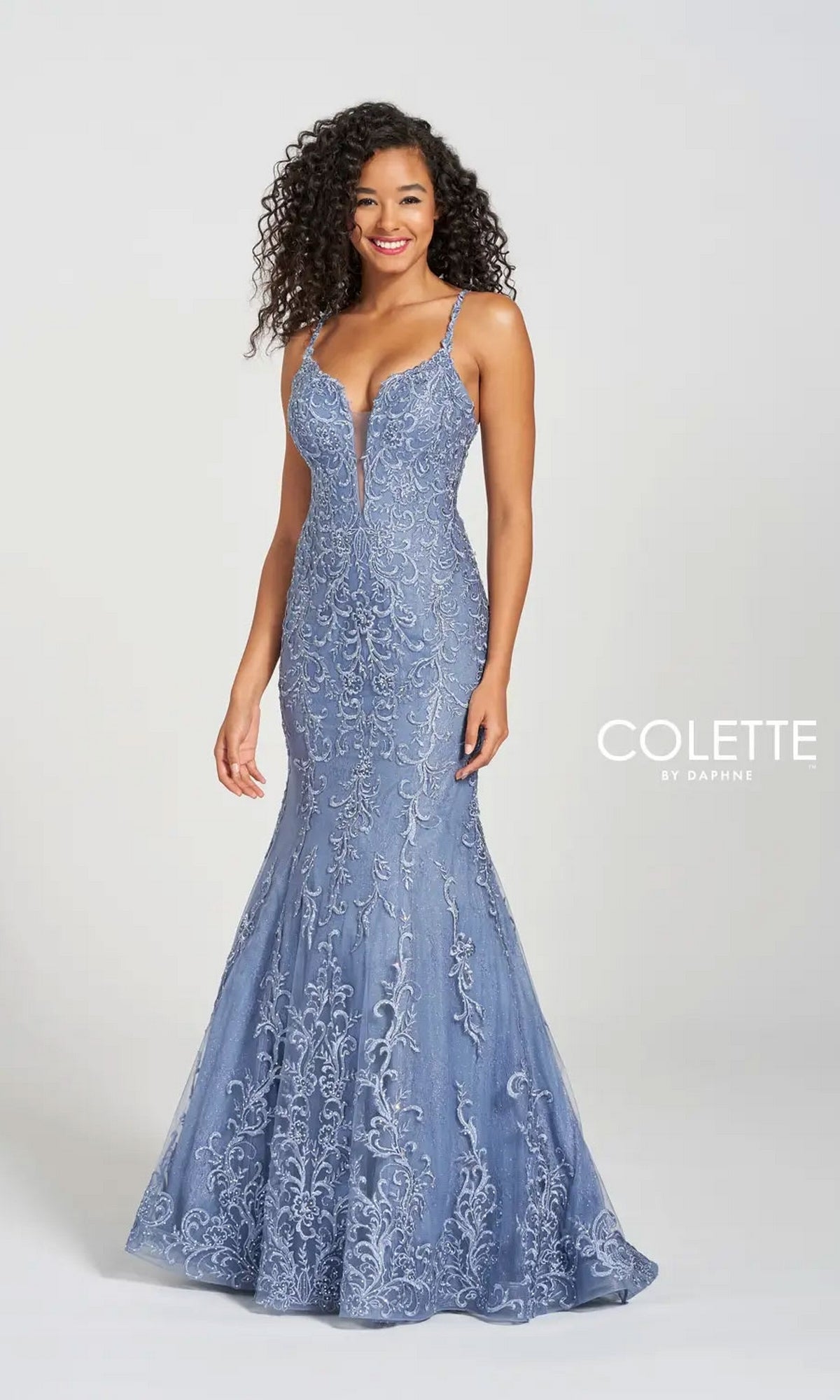 A woman wears the Long Prom Dress CL12220 by Colette—a fitted, sleeveless blue mermaid gown with a deep V neckline and vintage-inspired lace embroidery—standing against a plain light background for an elegant look.